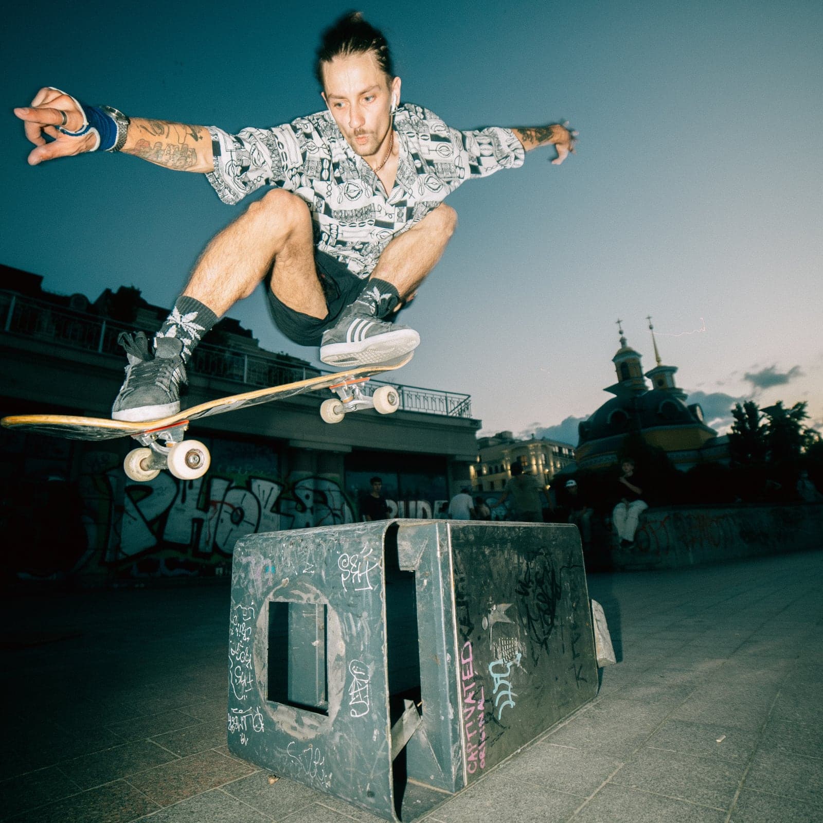 Skateboarder mid-trick over a graffiti-covered ledge on a city plaza at dusk