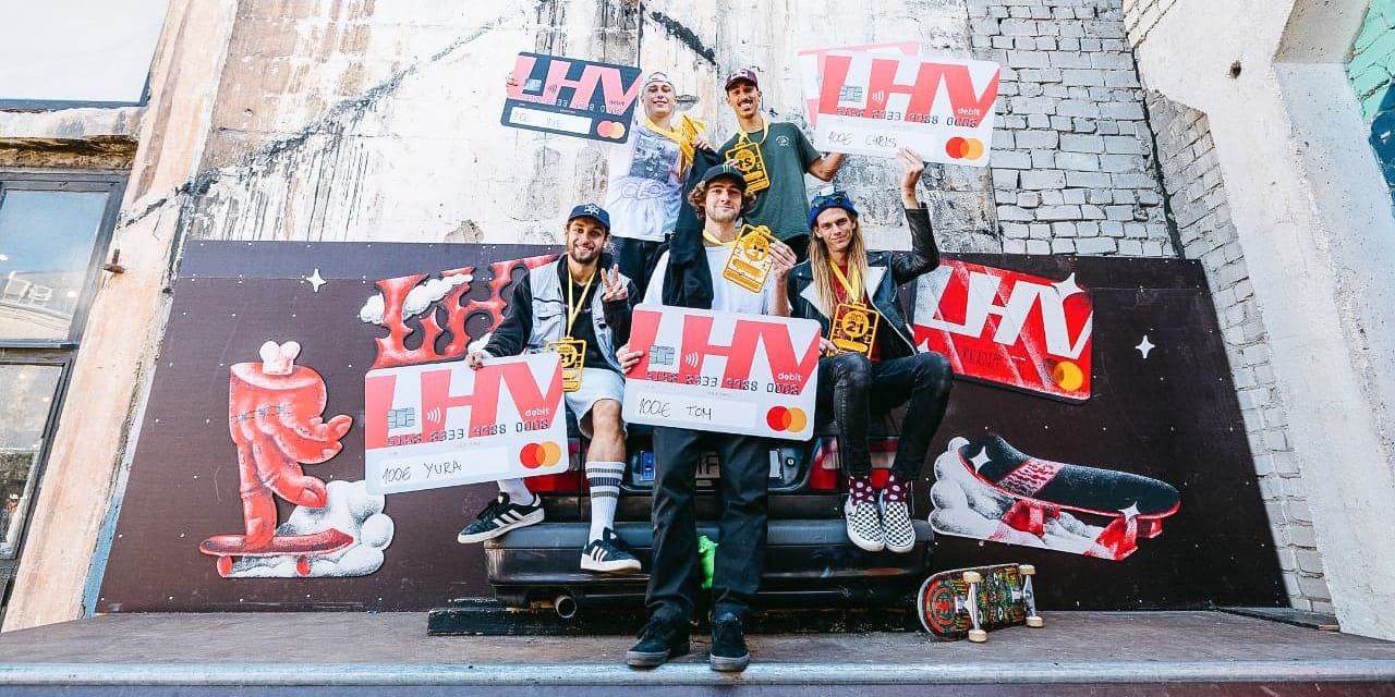 Skateboarders on a contest podium holding oversized prize cheques and medals in front of a painted mural
