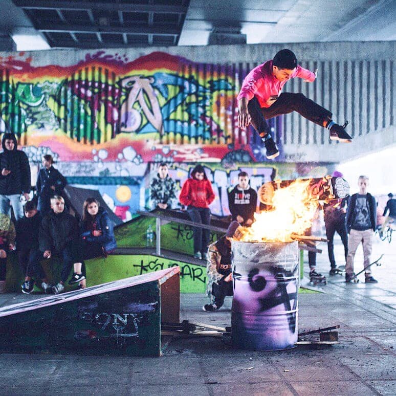 Skateboarder jumping over a burning barrel at a graffiti-covered spot under a bridge