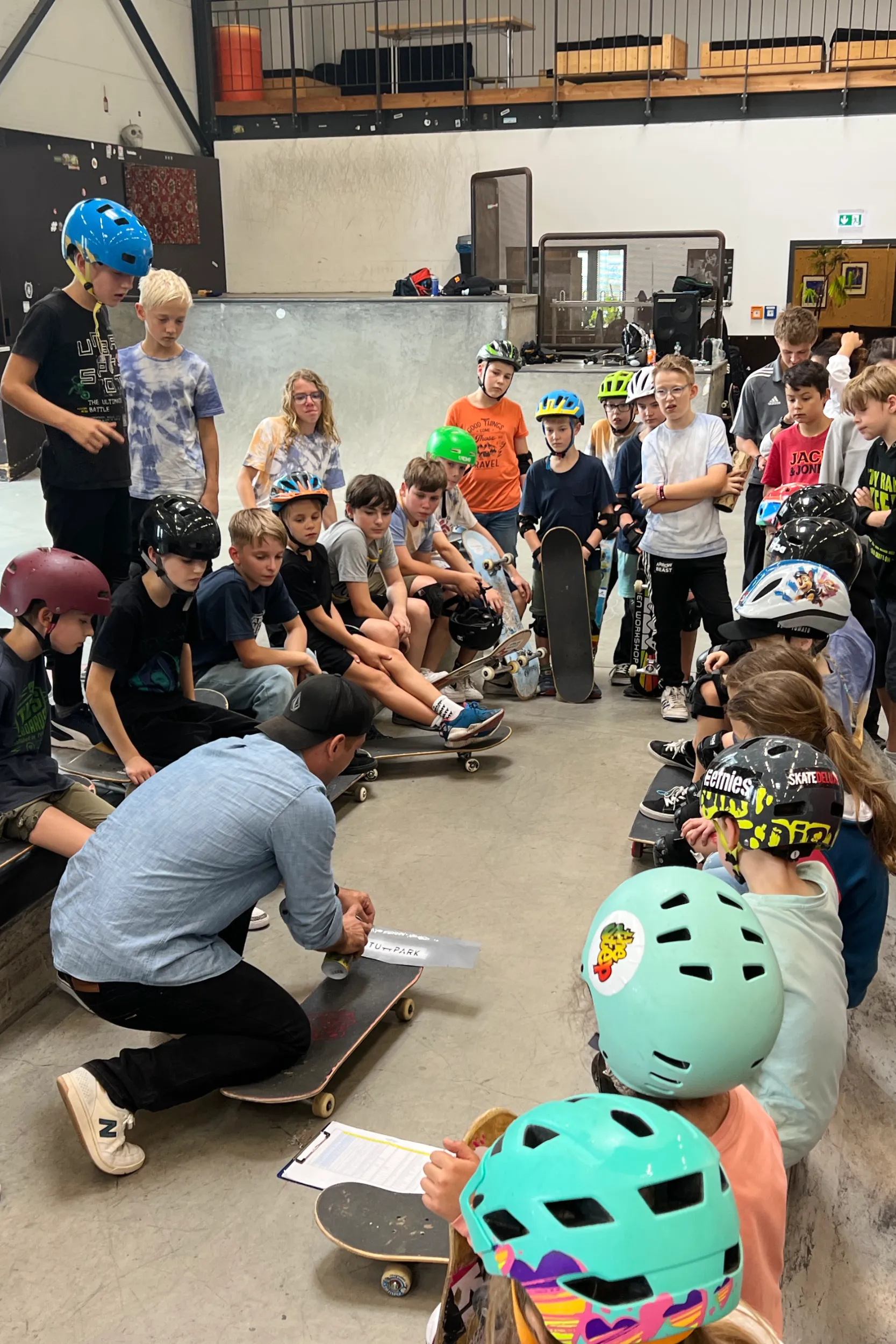 Coach griptaping a board in front of a seated circle of helmeted campers at an indoor skatepark