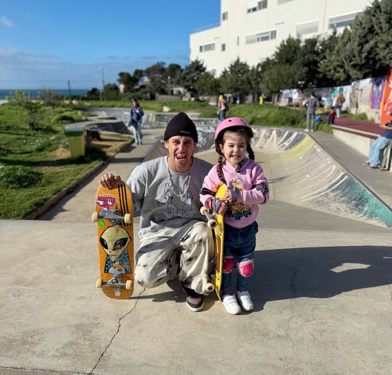 Skateboarding coach kneeling beside a young beginner in a pink helmet holding her first board at a seaside skatepark